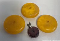 Three round amber butterscotch colored Bakelite disk beads showing surface texture and central hole, with dark amber pendant in silver-tone clasp for scale.