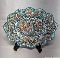 Front view of oval scalloped enamel dish with intricate hand painted floral and bird design on display stand