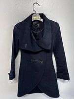 Front view of black Mackage Diana wool blend coat hanging on a hanger against a white wall, showing asymmetrical button closure and wide collar.