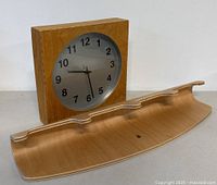 Wooden square wall clock with round metal face and wooden bentwood wall hook next to it.