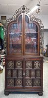 Front view of the full two-piece antique walnut display cabinet with curved top and metal patina metalwork.