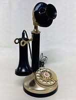Front view of vintage black and brass candlestick telephone with rotary dial, separate earpiece hanging on hook, and coiled cord.