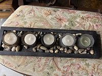 Photo of 5 glass tea light candle holders arranged on a wooden tray with decorative pebbles placed around them.