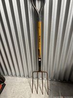 Photo of a garden fork with wooden handle and metal tines hanging against corrugated metal wall.