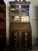 Front view of dark brown wooden secretary hutch showing closed glass paneled top doors with geometric mullions and three large drawers below with brass handles.