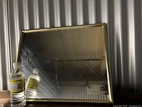 Gold framed rectangular mirror measuring 20 by 16 inches placed vertically, reflective surface with visible smudges, set against corrugated metal background.