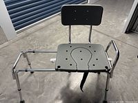 Front view of the transfer bench showing gray molded seat with an opening, armrests, backrest, adjustable legs, and safety strap hanging beneath the seat.