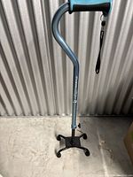 Photo of a blue walking cane with adjustable height and a four-prong base for support. Shows curved handle with wrist strap and the metallic body of the cane.