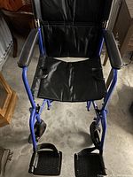 Front-facing photo of a blue wheelchair showing padded armrests, black seat and backrest, detachable footrests with straps, and storage pouch on the back