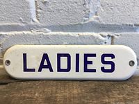 Front view of antique white enamel sign with 'LADIES' in blue lettering.
