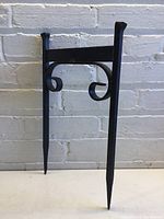 Front view of black metal boot scraper with two pointed legs and curled metal handles attached to a horizontal bar, standing vertically against a white brick wall.