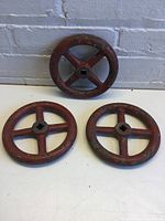 Three red-painted metal crank wheels with four spokes and square center holes laid on a white surface against a grey brick wall.