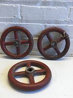 Three red metal crank wheels arranged against a white brick wall, showing overall shape, size, and condition.
