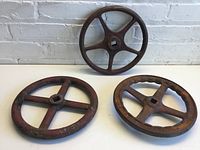 Three metal crank wheels of similar size and design laid on a white surface against a white brick wall.