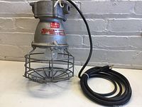 Metal industrial light fixture with attached power cord and metal cage surrounding the bulb area, shown from side angle.