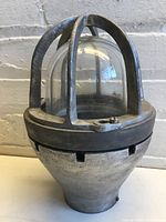 Photo of Crouse Hinds industrial light housing showing front view with protective metal cage and glass dome against a light brick wall background.