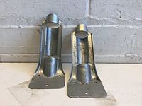 Two metal flag holders placed upright against a wall, showing front view of both holders, highlighting their curved design and mounting bases with holes.