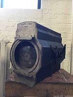 Large blue and silver vintage movie light by Kiegl Bros N.Y. with front lens visible, placed on wooden surface against brick wall background.