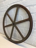 Large rusty metal wheel leaning against a brick wall, showing six spokes and central hole.