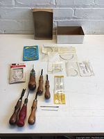 Full overview of all leatherworking tools and needles arranged on table with open box seen.