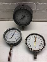 Three medium-sized round pressure gauges displayed on white surface with visible rust and wear.