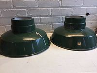 Pair of green enamel metal light shades shown side by side on a white surface with a brick background, showing color and shape