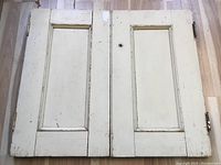 Full view of pair of off-white wooden shutters laid flat on floor, showing rectangular inset panels and hinges.