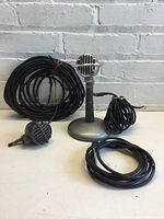 Front view of retro microphone on stand with extra microphone head and three cables coiled around it, set against white brick wall.