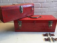 Two red metal tool boxes stacked with four metal cabinet hinges placed beside them