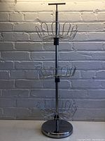 Full view of the triple basket with adjustable height, showing three metal wire baskets mounted on a chrome central pole with round metal base against a brick wall background.