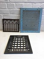 Photo of all three vent covers displayed against brick wall on table: blue rectangular vent cover with diamond mesh, black vent covers with circular lattice and arrow/arch design