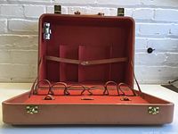 Opened vintage cocktail suitcase showing reddish-orange interior with bottle straps and compartments; one metal measuring cup present.