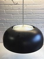 Front view of the black metal pendant light with white plastic concentric circular lens, hanging by a cord against a white brick wall.