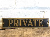 Close-up photo of the rectangular metal sign with brass letters spelling "PRIVATE" on a black background, mounted on a light-colored brick wall.