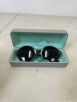 Black full rim oval sunglasses with gold accents resting inside a light blue-lined gray hard case.