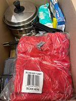 Box contents showing stackable stainless steel saucepans with lids, kitchen wraps, red fabric items in plastic packaging, and more household items inside a cardboard box.