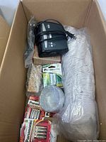 Top view of box showing Panasonic battery packs, black battery charger with power cord, roll of tape and bundle of white disposable cups.