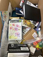 Photo showing a large cardboard box filled with various items including stationery, craft booklets, printer cartridge box, small toys in a bag, and other small boxed products.