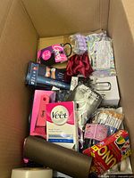 Top-down view of the box showing an assortment of women's accessories and personal care items including a pink Goody hairbrush, Veet wax strips, red scrunchie, black tweezers, and Uno cards.