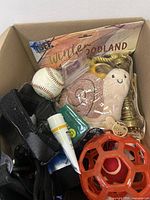 Box with assorted pet toys including plush snail toy, baseball, red plastic ball cage toy, and black strap.