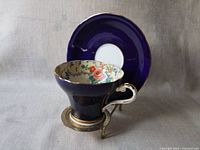Teacup in corset style with cobalt blue exterior, floral interior, gold rim, placed on metal stand with matching cobalt blue saucer behind it.