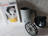 Silver cylindrical Bialetti milk frother with black handle, black lid with plunger, and detachable mesh press next to original packaging box marked Bialetti TUTTOCREMA.