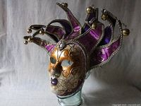 Front and side view of full face Venetian mask with jester headpiece in purple fabric and gold embroidery with metallic bells.
