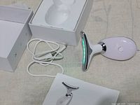 Facial and neck massager device with USB charging cable and packaging displayed on a fabric surface.