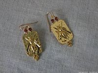 Pair of gold-tone metal earrings with embossed bee and pink rhinestones laid on gray fabric.