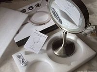 Close up of the mirror assembled with LED light off, clear view of magnifying mirror side, USB cable, instruction manual and original box.