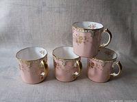 Four Royal Crown Derby demitasse/espresso cups, pink with ornate gold floral pattern, displayed grouped with one cup stacked atop another.
