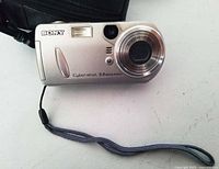 Front view of the silver Sony Cyber-Shot digital camera, showing 5.0 megapixels label, lens, flash, and wrist strap attached.