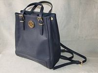 Front angled view showing navy blue leather handbag backpack with gold-tone logo emblem and handles.