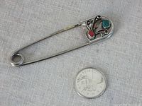 Top view of vintage safety pin brooch next to a quarter for size reference, showing the turquoise and red stones and detailed metalwork.